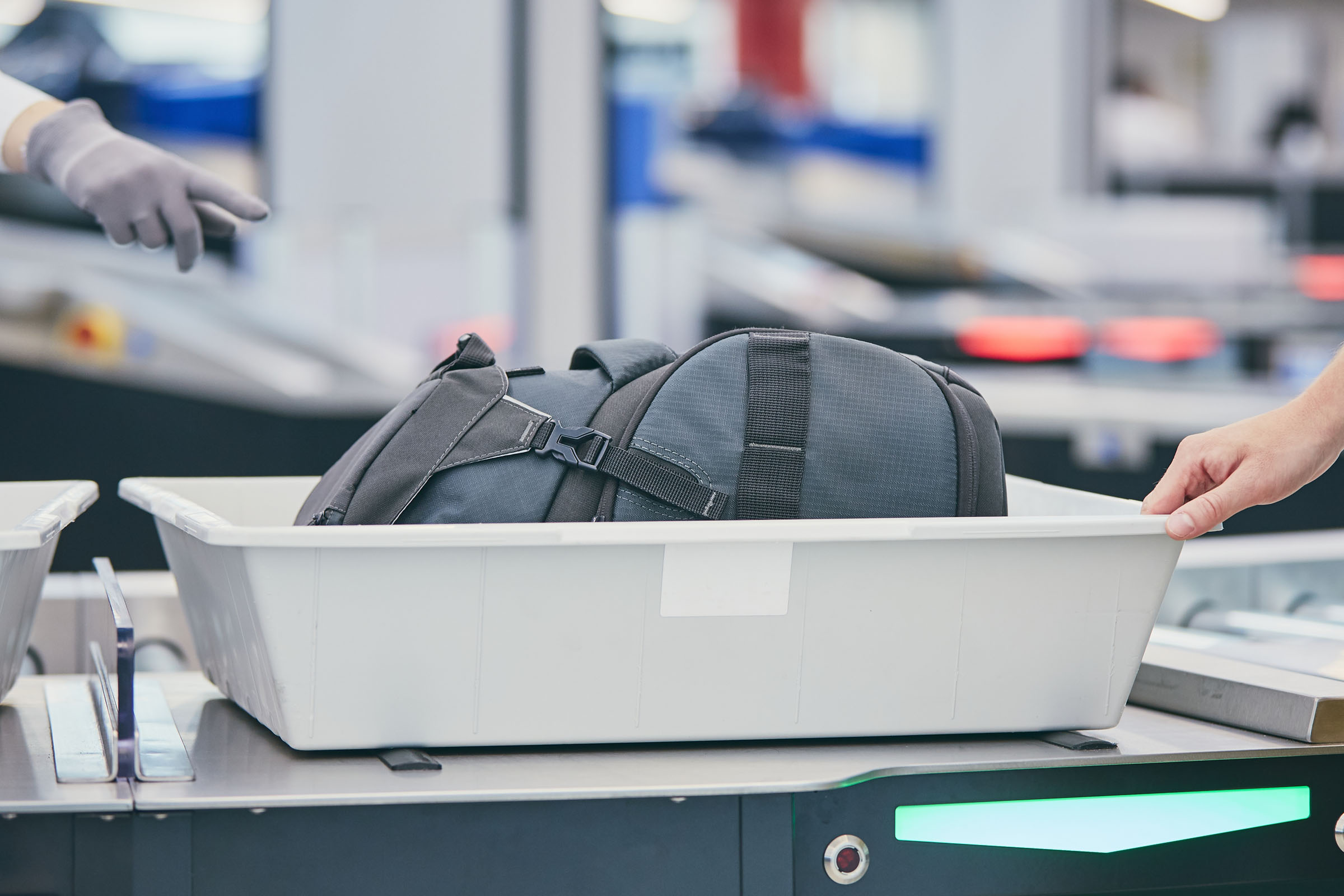 A gloved hand points to a bag in a plastic bin that another hand is pulling toward a scanner.